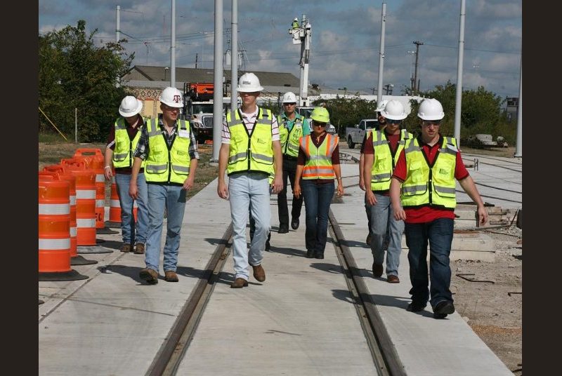 Students wearing construction hats, safety vests and goggles walk along a train track