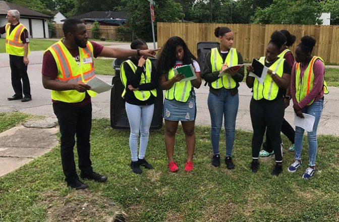 A man wearing a safety vest instructs a group of students wearing safety vests and working in a neighborhood