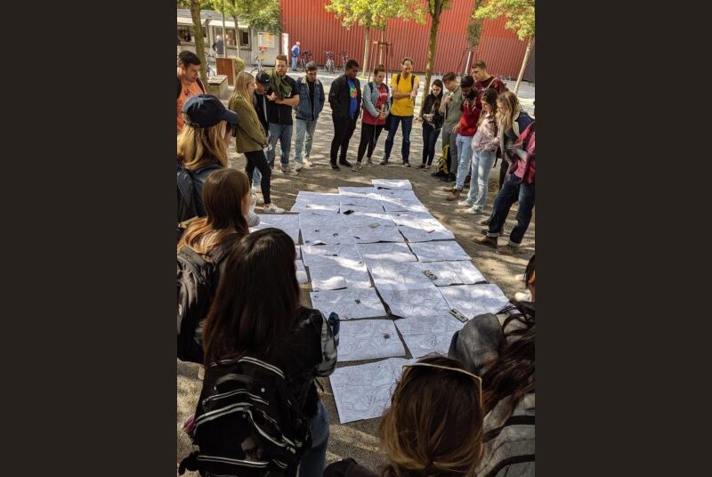 Shaded by trees, students gather in a circle to view several land development plans on the ground