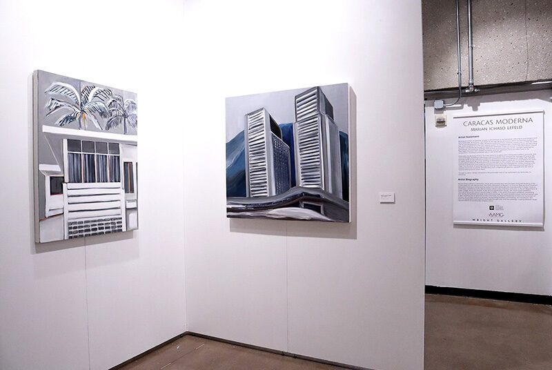A view of the gallery with two walls with two artworks on display and a large sign with text in the background. On the left, there is a black and white painting of a beachside house with palm trees in the background. On the right, there is a black and white painting of two skyscraper buildings placed on a blue and gray background.
