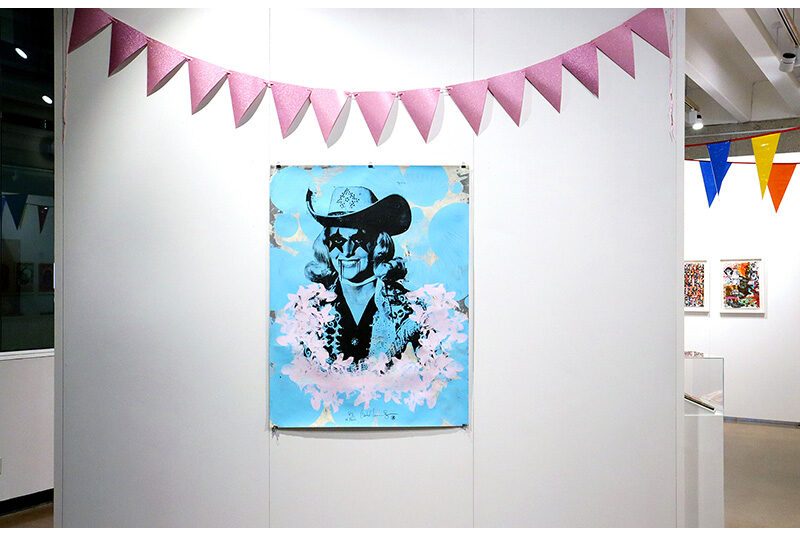 On a white wall, a large portrait of a woman is centered. The portrait has a blue background and the woman wears a cowboy hat with star shaped face paint around her eyes. Above the artwork, a pink banner hangs on the wall.
