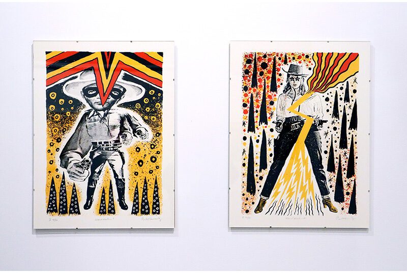 Two prints are installed on a white wall. Both prints have black, white, red and yellow. The image on the left features an abstracted cowboy with a pistol. The image on the right depicts a cowgirl with a pistol. In both images, the figures have wide stances and the pistol points at the viewer. Abstract shapes and color throughout the backgrounds.