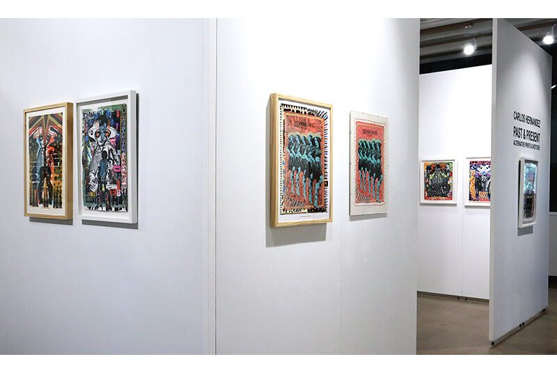 Several white walls recede into the distance of the art gallery with small artworks on the walls.On the right, there is black text on the wall above an artwork that says “Carlos Hernandez Past & Present: Alternative Prints & Sketches.”