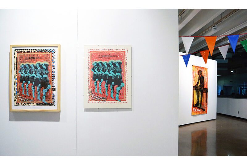 Two small posters on the left side depict similar imagery of cowgirls engaged in a line dance; text reads Marfa Texas. In the background, a large orange artwork is on the wall. The imagery is a skeleton playing an electric piano. A party banner of colorful triangles hangs between the walls.