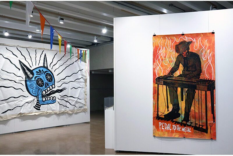 A large tapestry on the left has a painted blue skull with horns and a large tongue. On the right, a large artwork depicting a skeleton dressed in Western outfit, seated and playing an electric piano.