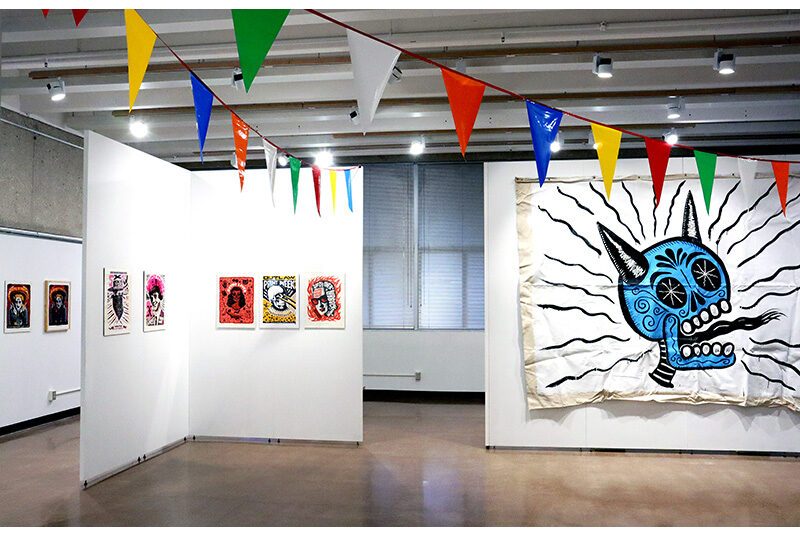 An expansive view of an art gallery with many of Carlos Hernandez’ artworks hanging on the wall. Several small prints are installed on walls on the left side of the image. On the right, a large tapestry hangs on the wall. Towards the top, two rows of party banners with colorful triangles hang from the walls.