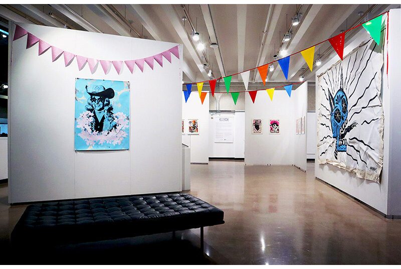 On the left, a large leather bench is centered in front of a wall with a blue portrait of a woman. On the right, a series of walls with artwork recedes into the distance of the art gallery. Towards the top of the walls, rows of party banners with colorful triangles are suspended throughout the art gallery.