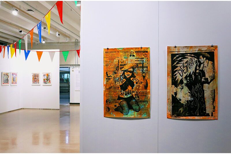 In the foreground, a set of two orange artworks hang on white walls. The artwork on the left depicts a woman holding two pistols in the air. On the right, the image depicts a man wearing Western attire and singing towards a crowd of people. The lyrics say “destroy” inside a speech bubble. On the left of the photograph, two small artworks hang on the wall. Towards the top of the walls, rows of party banners with colorful triangles are suspended throughout the art gallery.