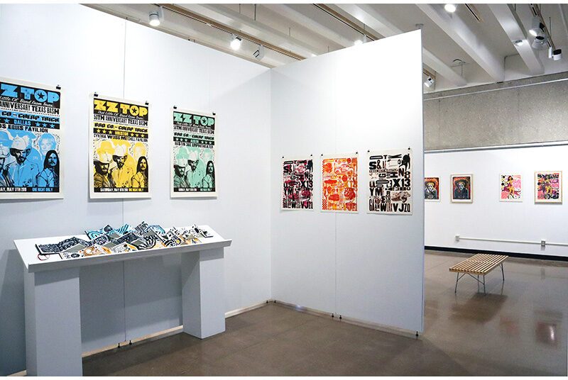 A room with long shelf holding accordion style sketchbooks with the artist’s drawings. Above the self, 3 posters promoting the musical group, ZZ Top. In the center of the photograph, a set of 3 prints on the walls features various text created with different shaped letters. In the background, a wooden bench is in front of 4 posters on the wall.