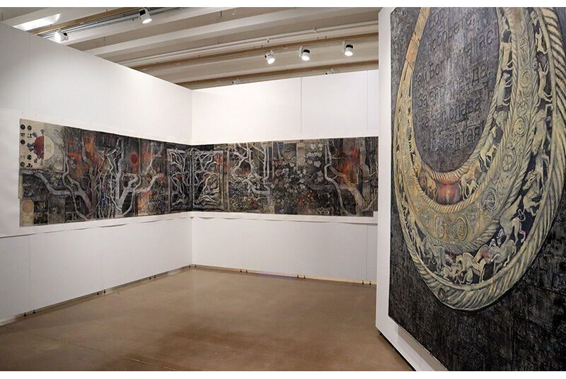 A view of two artworks. The artwork on the left spans most of the 2 walls it is mounted on. The artwork on the right features a dark background and a neutral horseshoe shape design.