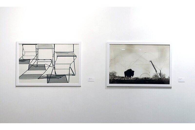 A view of 2 black and white photographs in white picture frames. On the left, there is a photograph of 3 chairs with shadows. On the right, there is a photograph of an open field with a structure on the left side of the image.