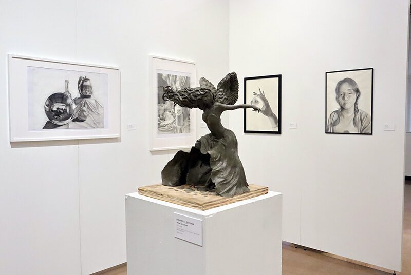 A figural statue on a pedestal sits in front of 4 framed graphite drawings in the background. The figural sculpture is in the center with her arms spread wide and hair flowing in the wind. There are 2 still life drawings on the left, 1 drawing of a hand holding mirror, and 1 portrait of a young woman.