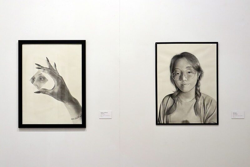A view of 2 graphite drawings. On the left there is a drawing of a hand with long nails holding a square mirror with eyes reflected inside. On the right there is a self portrait of a young woman.