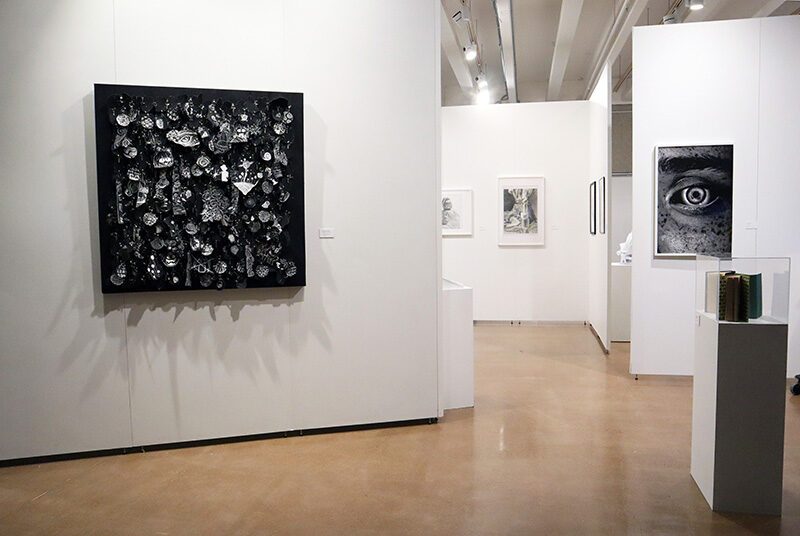 On the left, on the wall there is a black and white abstract artwork fabricated from multiple student’s drawings. On the right are books on top of a pedestal with a photograph of a large eye. In the background are framed artworks on the wall.