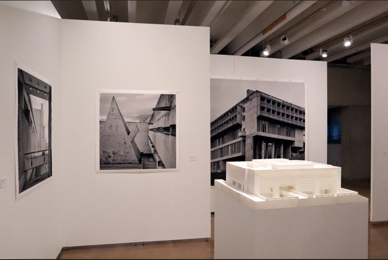 A 3D-printed model of Texas A&M’s Langford Building on a white pedestal with Leonid Formansky’s black and white photographs on a white wall behind it.