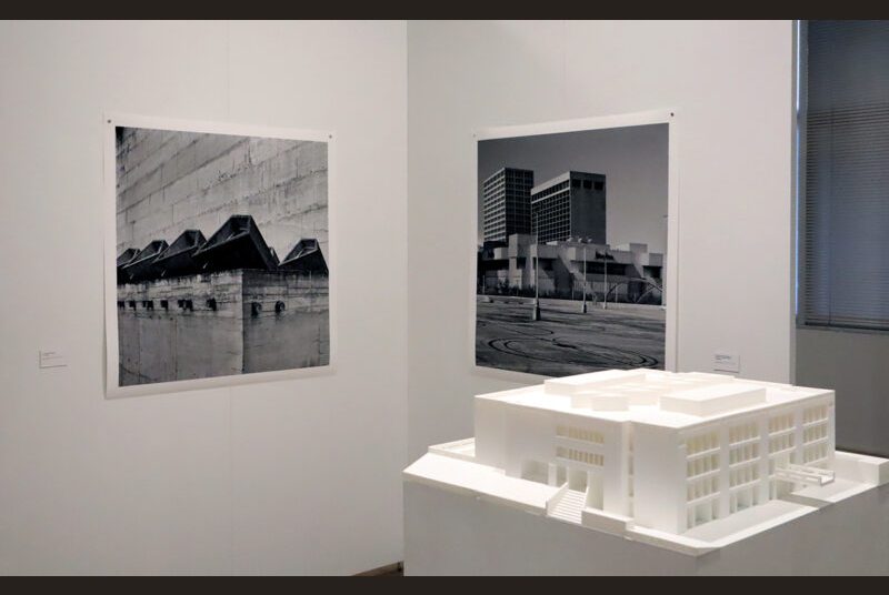 A 3D-printed model of Texas A&M’s Langford Building on a white pedestal with “La Tourette Convent 3” and black and white photos of architecture such as “Neiman Marcus Houston TX” behind it on a white wall.