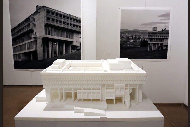 A 3D-printed model of Boston City Hall on a white pillar with a print of Leonid Formansky’s “La Tourette Convent #0” and “La Tourette Convent #6” on a white wall behind it.