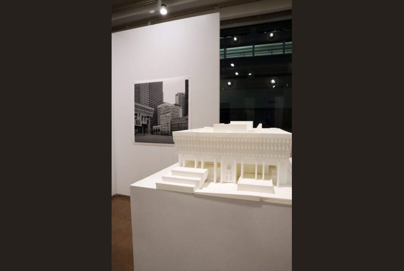 A 3D-printed model of Texas A&M’s Langford Building on a white pillar with a print of Leonid Formansky’s “Boston City Hall” on a white wall behind it.