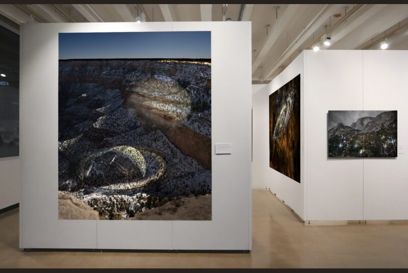 A large-scale photograph of Grand Canyon with projections of Saturn and Jupiter on it, installed on a white wall. In the background, there are two additional photographs installed on the gallery walls.