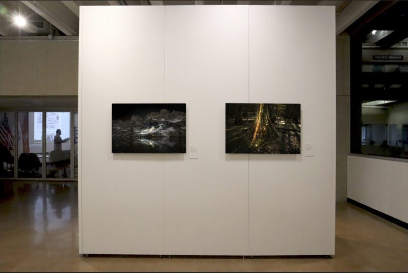 Two small photographs of landscape with space imagery are installed on a white wall. In the background, there is an adjacent lecture room with a man standing with hands out while presenting a lecture.