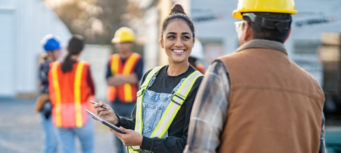 A female foreman reviews plans electronically on a tablet with a crew member as they review the site tasks together. Other crew members can be seen conversing in the background.