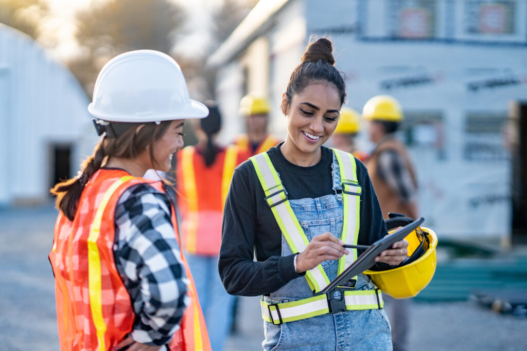 A female foreman reviews plans electronically on a tablet with a crew member as they review the site tasks together. Other crew members can be seen conversing in the background.