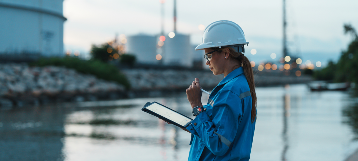 Female petroleum oil refinery engineer working late with tablet to check safety quality control of oil and gas plant at dusk.