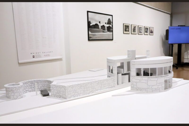 A white small scale model of a round house is in front of photos and a sign on the wall. A tv monitor in the background on the right.