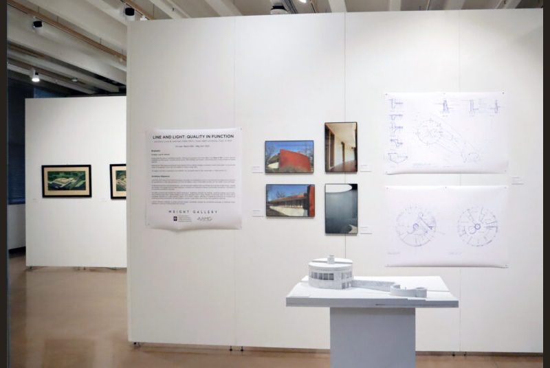 A white small scale model of a round house is in front of photos and a sign on the wall. In the background there is a wall with 2 paintings on it.