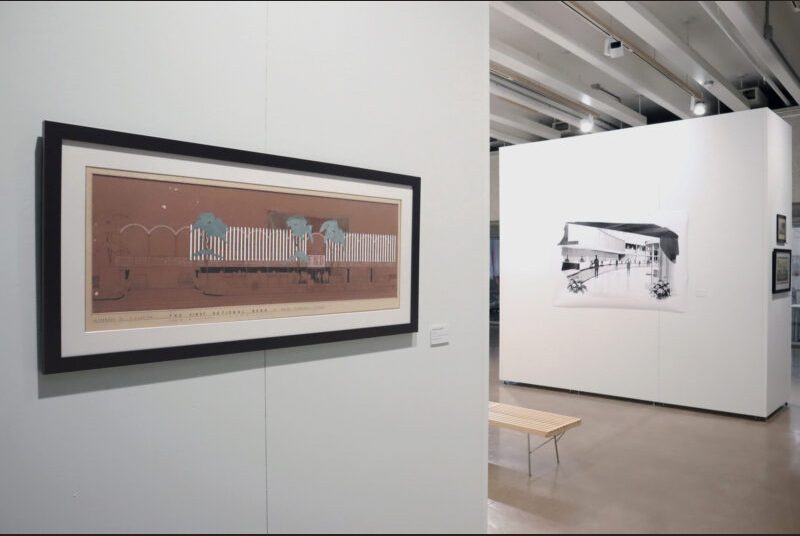 A painted elevation of a bank on brown paper is framed with black and white molding. In the background, a large black and white painting of a bank is on the wall behind a wooden bench.