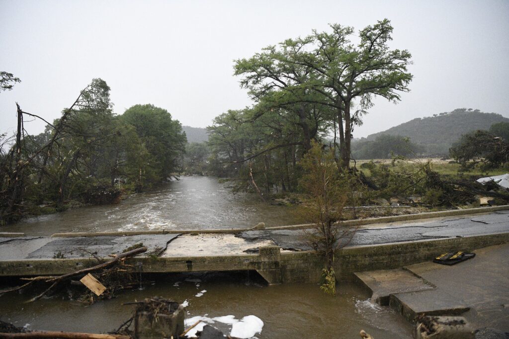 An old bridge stands surrounded by high floodwaters, with broken tree branches and debris scattered along the swollen riverbanks.
