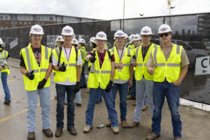 Six students wearing hard hats and high-visibility vests pose in front of a gated active construction site.