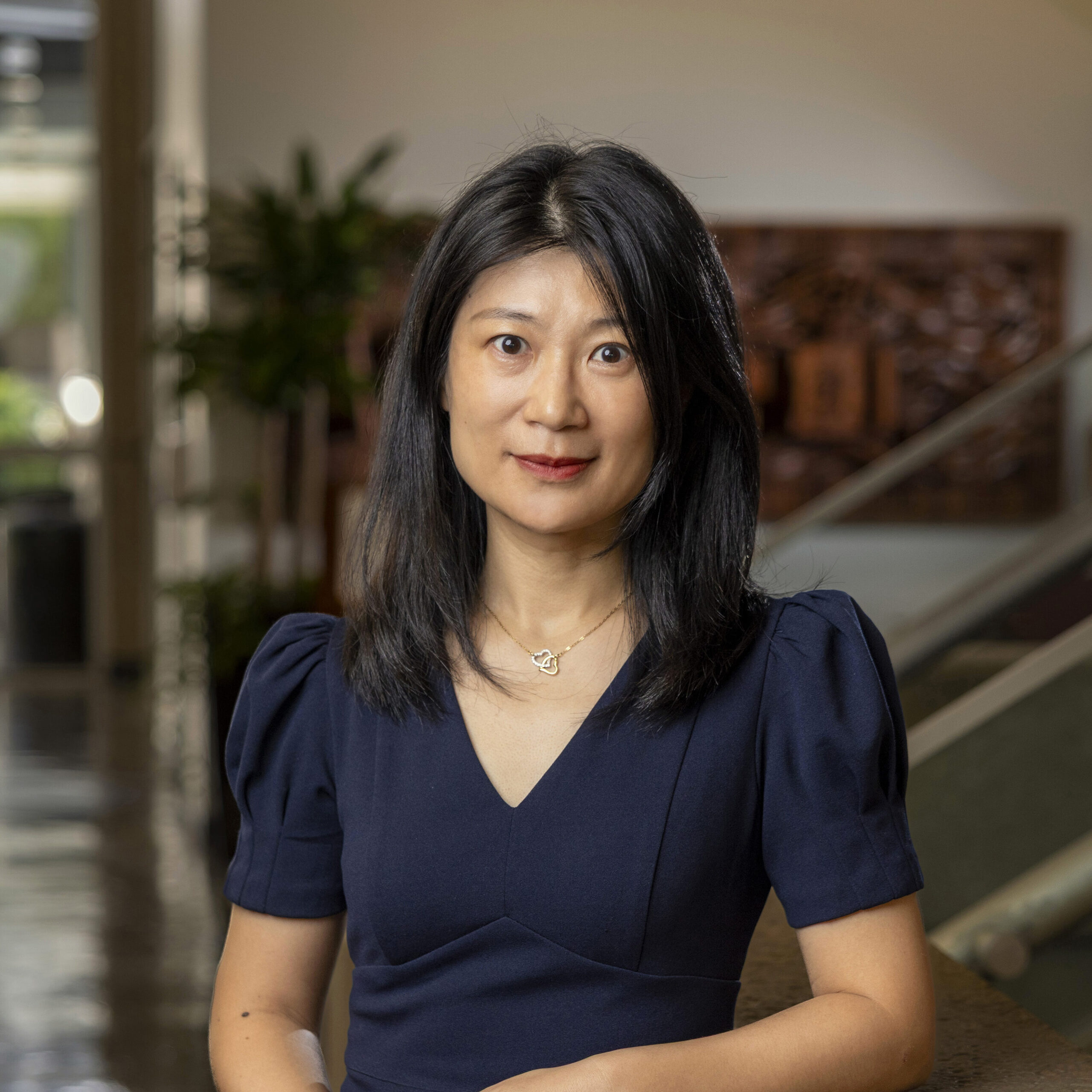 Name: Dean Ivy Hu,A woman with black hair wearing a navy dress stands indoors with a calm expression. Background includes modern architecture and green plants.