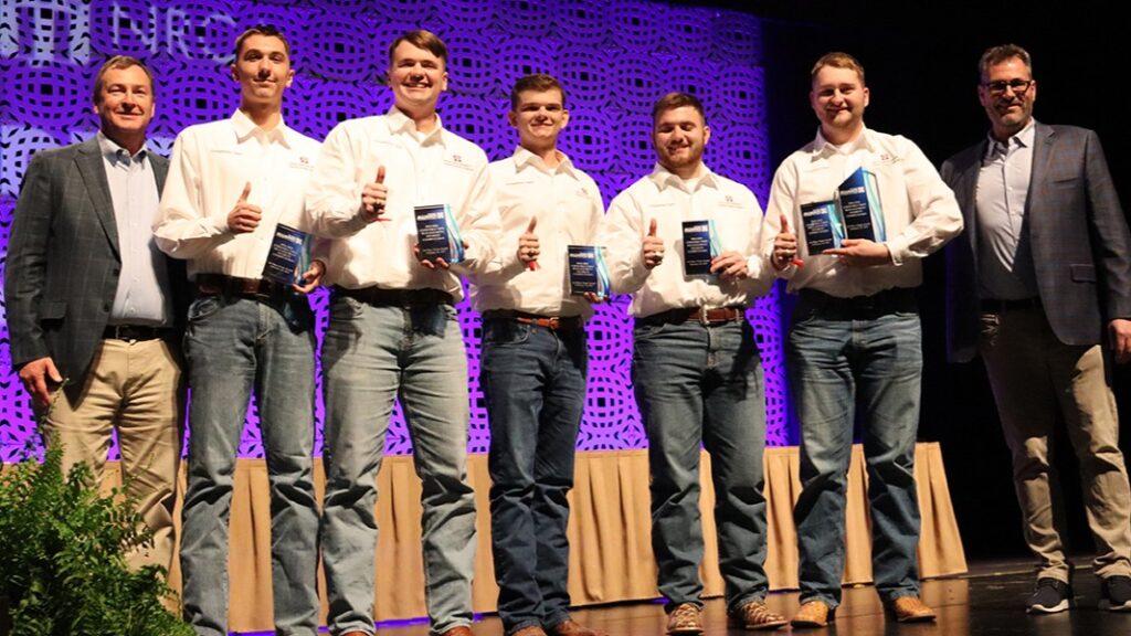 Five students holding trophies, standing with two faculty members on a stage.